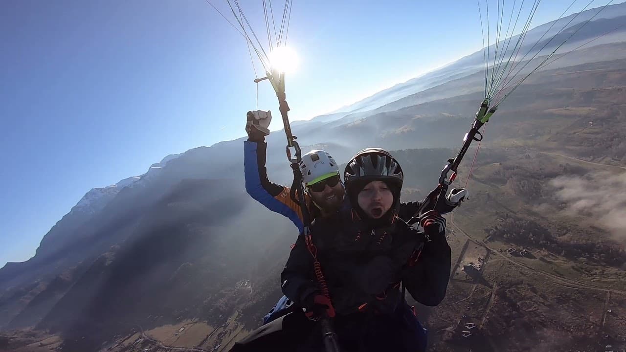 Pilot and passenger on a tandem flight, with the sun above the wing and mountain ridges behind