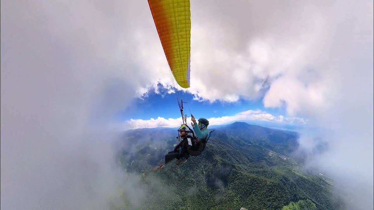 Tandem flight among the clouds, with the yellow-orange wing above and forested mountains below